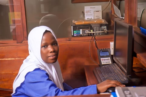 Young student in white hijab and blue uniform working at a computer desk in a classroom setting