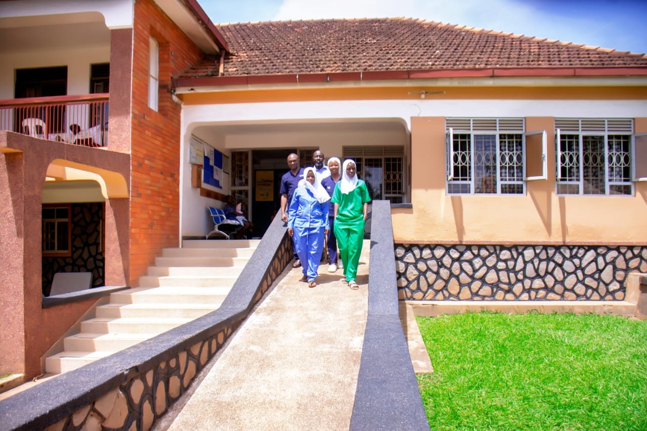 Healthcare workers in blue and green uniforms walking on a paved pathway toward a brick medical facility with white trim