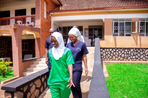 Two female healthcare workers in green and blue scrubs with white headscarves walking outside a medical facility courtyard