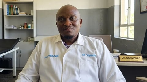 Professional man in white lab coat seated at desk in office with shelves and nameplate visible